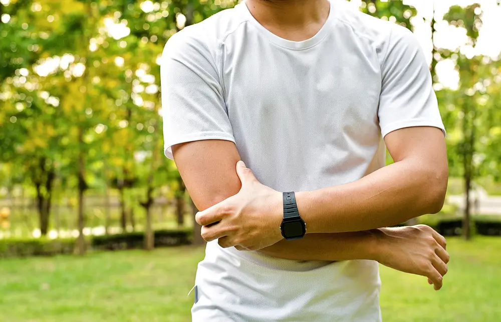 man holding bicep, trees and grass blurred in the background