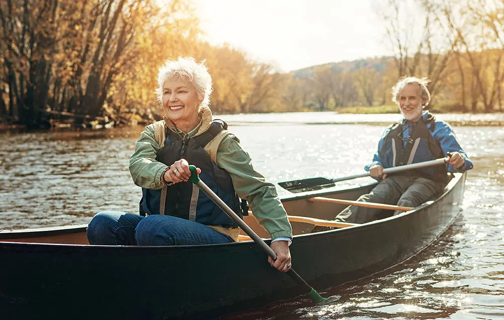 Two people smiling and paddling a canoe on a calm river surrounded by autumn trees. The sun is shining, creating a warm, golden glow.